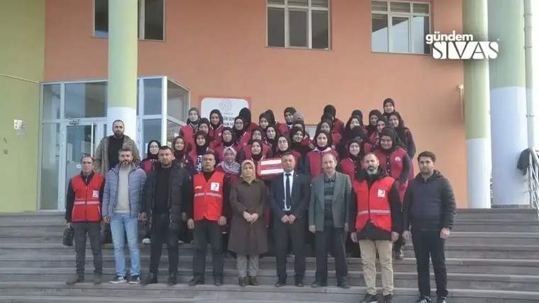 Bir grup öğrenci ve yetişkin, okul önünde poz veriyor; bazıları kırmızı yelek giymiş.