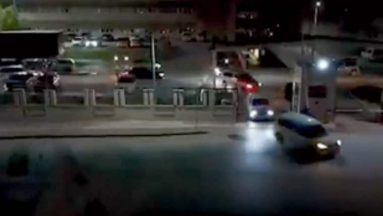 Gece saatlerinde, aydınlatılmış bir kavşakta ilerleyen araçlar ve yol kenarında bekleyen otomobiller görülüyor.