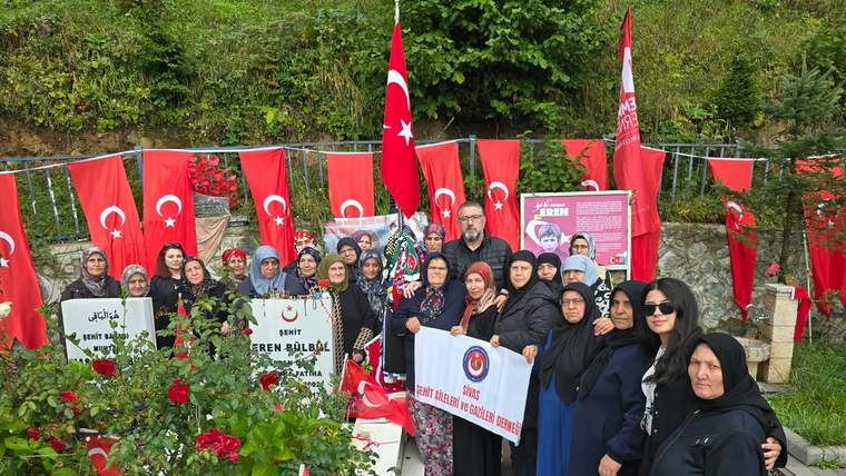 Türk bayrakları ve çiçeklerle süslenmiş alanda, üzerinde Eren Bülbül yazan pankartla duran bir grup kişi