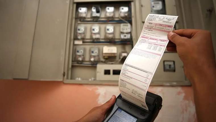 Bir kişi, elektrik sayacının önünde faturayı tutuyor.