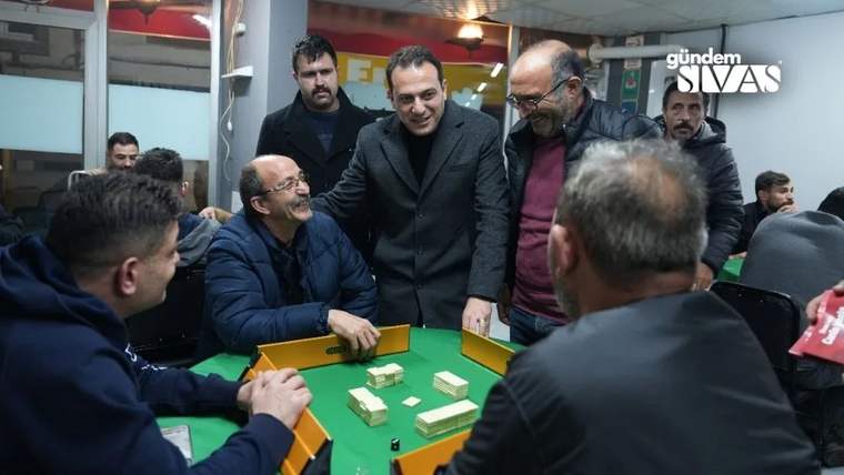Bir grup erkek, bir masanın etrafında oturmuş, oyun oynuyor ve gülümseyerek sohbet ediyor.