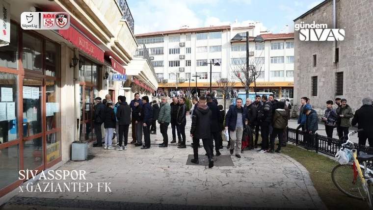 Bir dükkânın önünde, kaldırım boyunca uzanan sırada bekleyen kalabalık ve çevrede duran kişiler görülüyor.