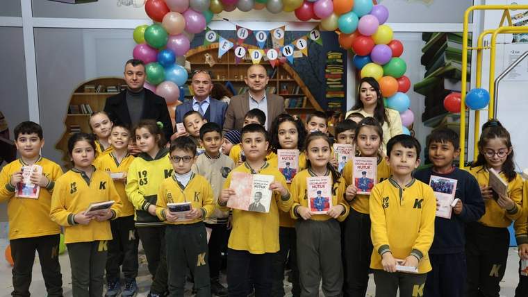 Kitaplıkların önünde sarı tişörtlü çocuklar, dört yetişkin ve renkli balonlarla süslenmiş bir alan görülüyor.