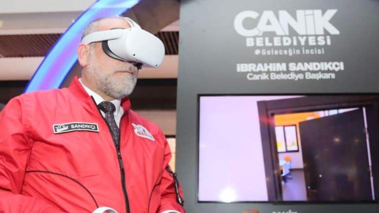 Kırmızı montlu bir kişi sanal gerçeklik gözlüğü takarken, yanında Canik Belediyesi yazılı ekran bulunuyor.