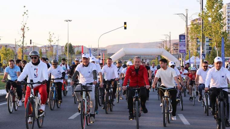Trafiğe kapalı geniş bir caddede çok sayıda bisikletli ilerliyor; arkada trafik ışıkları ve modern yapılar görülüyor.