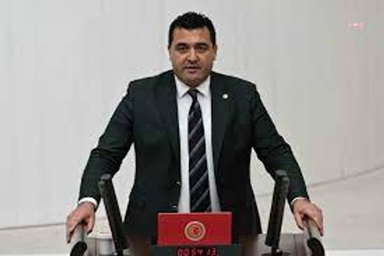 Takım elbiseli bir kişi, kırmızı amblemli kürsüde iki mikrofon arasında konuşuyor.
