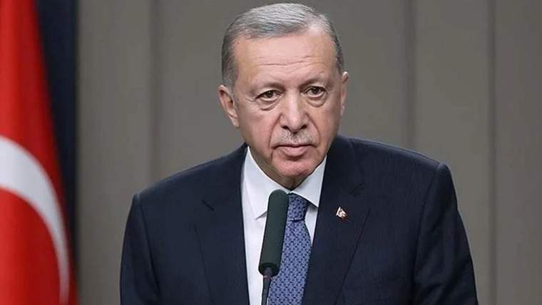 Türk bayrağının önünde, kürsüde mikrofonla konuşan koyu renk takım elbiseli bir kişi görülüyor.