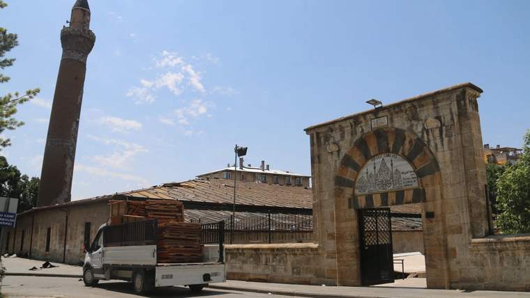 Taş kemerli giriş ve minaresi görünen tarihi caminin önünde küçük bir kamyonet duruyor.