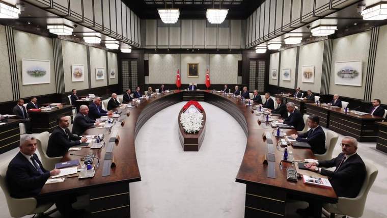 U biçimli masalarda oturan çok sayıda kişi, ortada çiçek aranjmanı ve duvarda iki Türk bayrağı görülüyor.