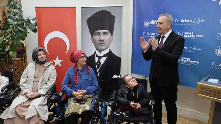 Engelli bireylerin bulunduğu bir etkinlikte, bir kişi konuşma yapıyor. Arkada Türk bayrağı ve Atatürk posteri var.
