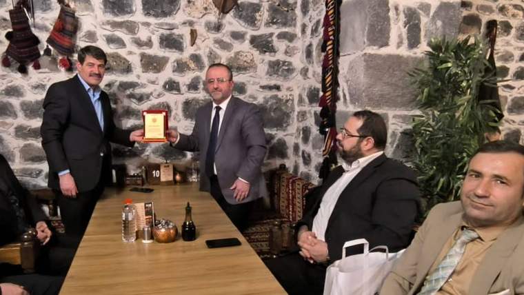Taş duvarlı bir salonda dört kişi oturuyor; ayakta duran iki kişiden biri diğerine plaket uzatıyor.