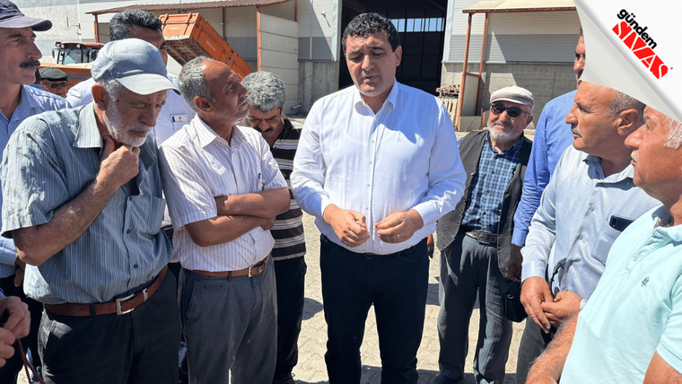 Bir grup insan, bir kişiyle sohbet ediyor; arka planda tarım alanı ve yapı görünüyor.