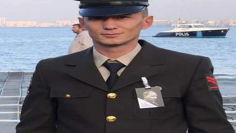 Deniz kıyısında üniformalı ve kep takan bir adam, göğsünde portreli kartla duruyor; arkada polis teknesi görülüyor.