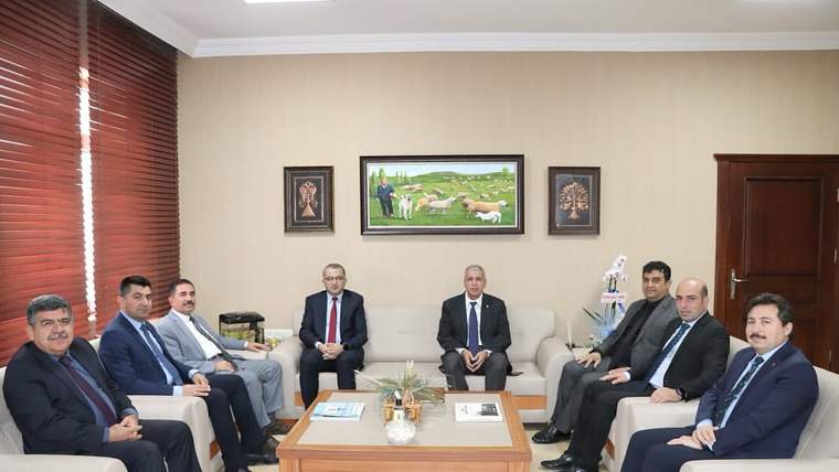 Bir grup erkek, ofis ortamında bir araya gelerek oturuyor. Arkada bir tablo ve bitkiler var.