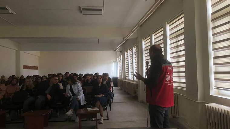 Sınıfta kırmızı yelekli bir kişi, karşısında oturan öğrenci grubuna konuşurken görülüyor.