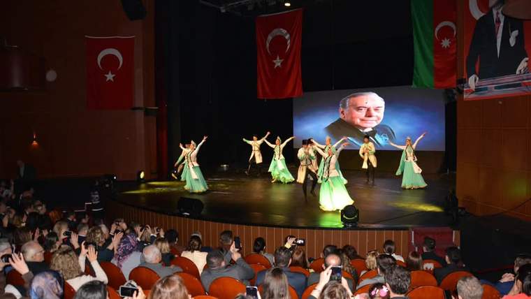 Sahnede geleneksel kıyafetli dansçılar gösteri yaparken arkada Türk ve Azerbaycan bayrakları görülüyor.