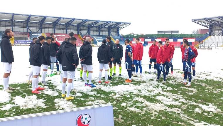 Karlı zemindeki futbol sahasında, kırmızı ve siyah montlu kişiler ile futbolcular bir arada duruyor.