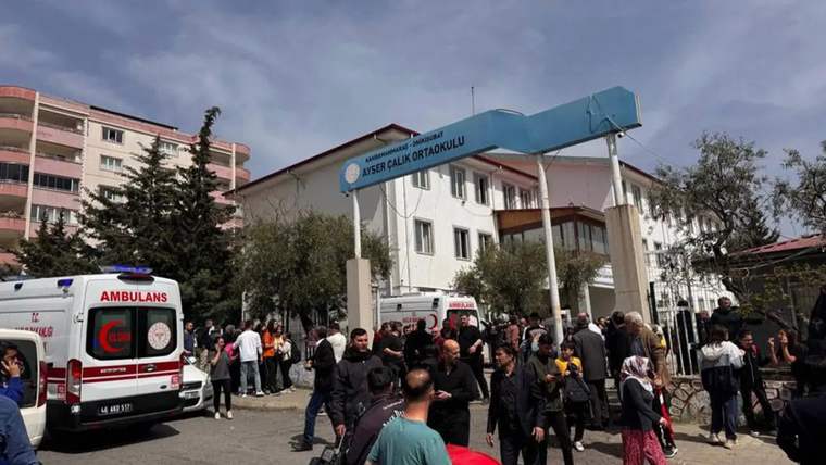 Kalabalık bir grup insan, bir okulun önünde toplanmış. Arka planda okul binası ve ambulanslar görünüyor.