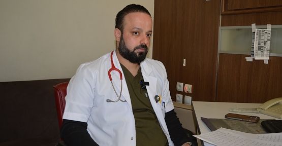 Beyaz önlüklü, stetoskop takan sakallı bir doktor, masa ve bilgisayarın bulunduğu odada oturuyor.