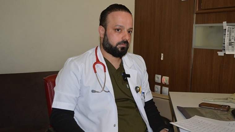 Beyaz önlüklü, stetoskop takan sakallı bir doktor, masa ve bilgisayarın bulunduğu odada oturuyor.