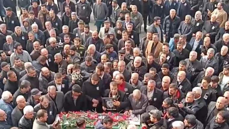 Çok sayıda kişinin çevrelediği tabutun üzerinde kırmızı çiçekler bulunuyor; kalabalık açık alanda toplanmış.