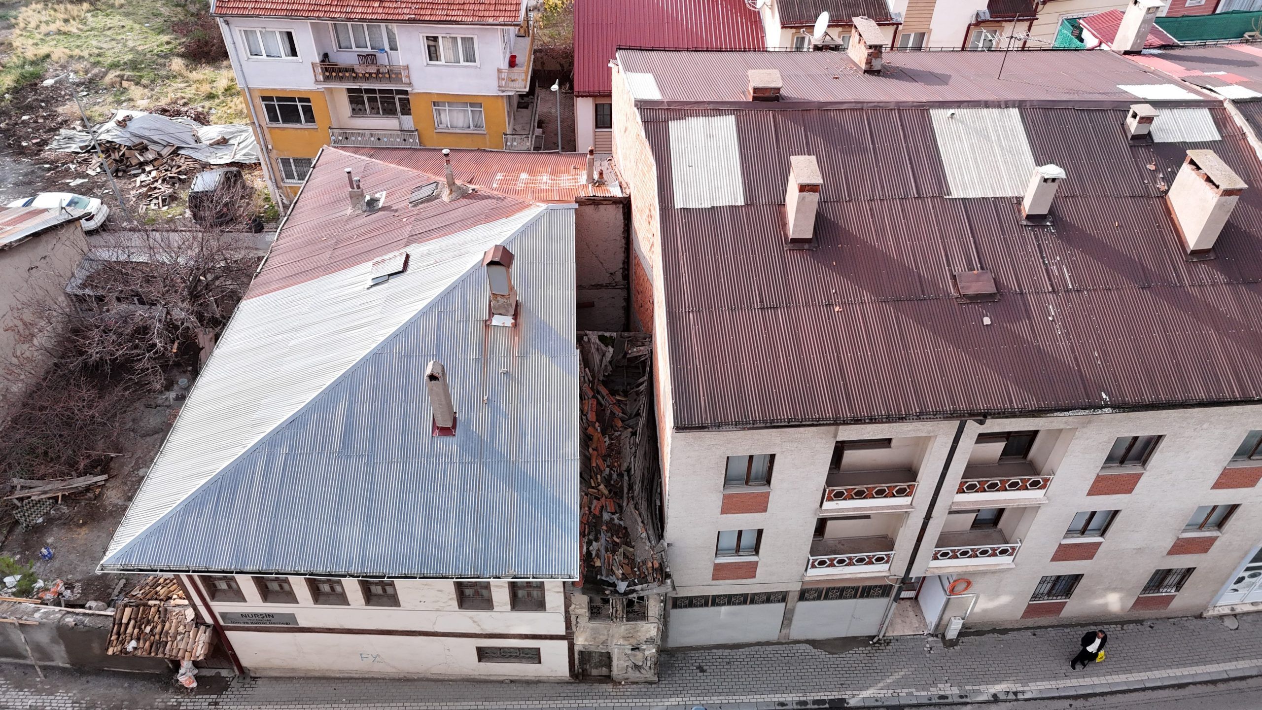 İki katlı bir metruk bina, yanındaki binaların arasında yer alıyor. Bina, mavi çatılı ve yanındaki binalarla yakın mesafede.
