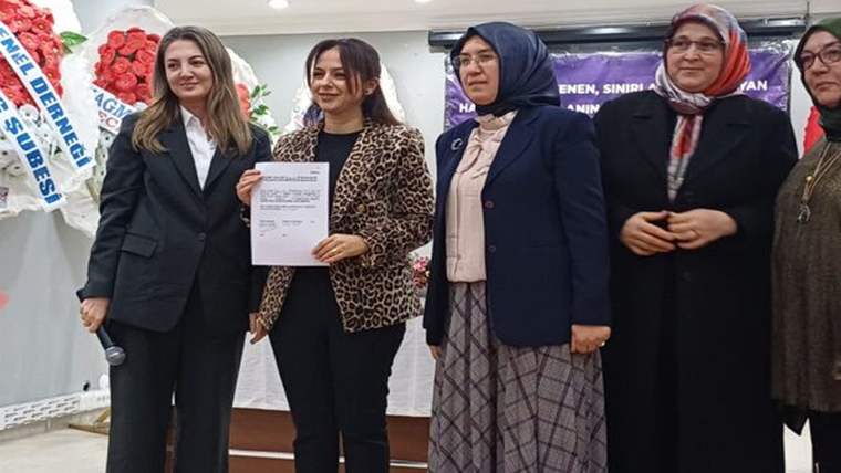 Kapalı salonda dört kadın yan yana duruyor; ortadaki kadın elinde belge, soldaki kadın mikrofon tutuyor.