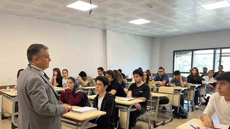 Sınıfta sıralarında oturan öğrenciler ve önlerinde ayakta duran takım elbiseli bir kişi görülüyor.