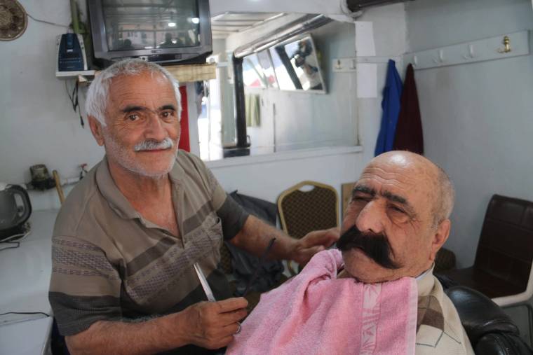 Berber, koltuğunda oturan ve pembe örtü takılmış müşterisinin saçını makasla kesiyor.