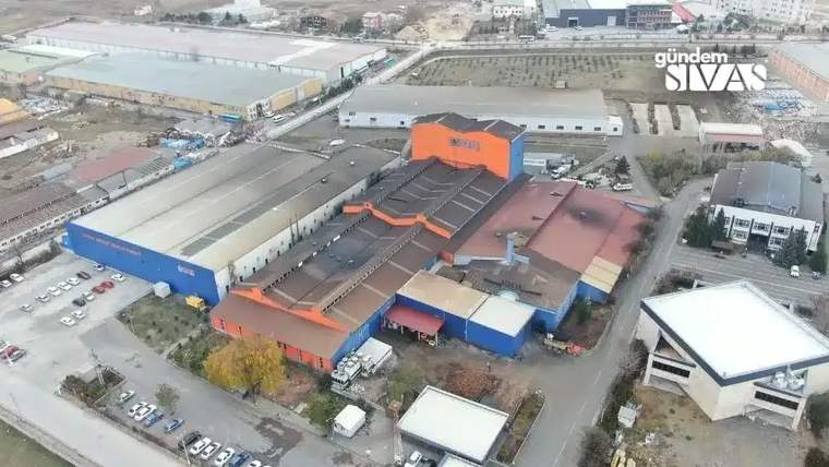 Bir sanayi tesisi, çevresinde araçlar ve ağaçlar ile birlikte görünmekte.