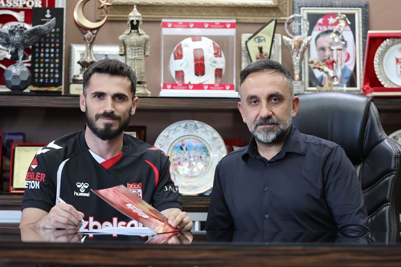 Siyah forma giymiş bir futbolcu, masada bir belge imzalarken yanında bir kişi oturuyor.