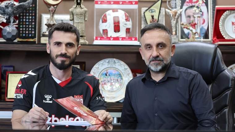 Siyah forma giymiş bir futbolcu, masada bir belge imzalarken yanında bir kişi oturuyor.