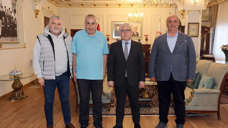 Dört kişi, bir oturma odasında yan yana duruyor; arka planda lamba ve fotoğraflar var.
