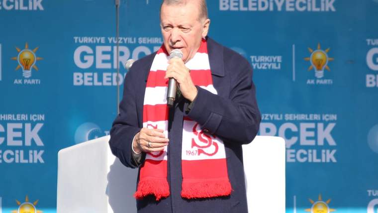 Mikrofonla konuşan bir kişi, kırmızı-beyaz atkısıyla mavi ve yazılı bir sahne önünde duruyor.