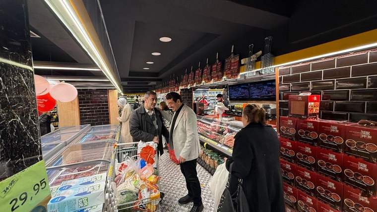 Market reyonunda üç kişi alışveriş arabaları ve ürünlerle birlikte dururken arka planda et tezgâhı görülüyor.