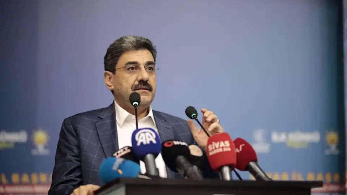 Kürsüde konuşan bıyıklı bir kişi, önündeki çok sayıda logolu mikrofon arasında elini kaldırıyor.