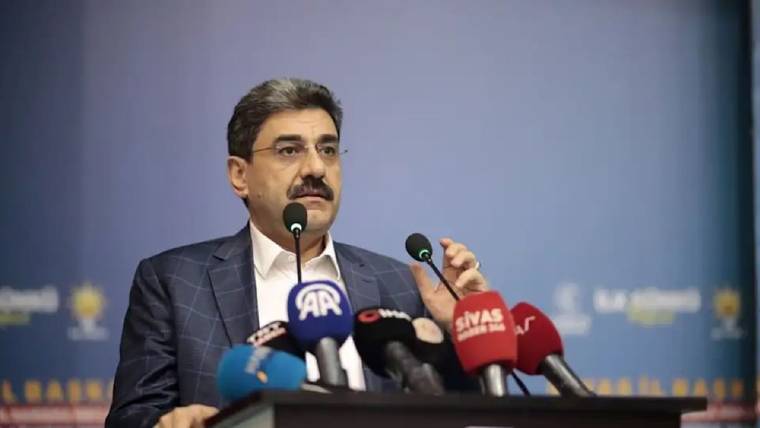 Kürsüde konuşan bıyıklı bir kişi, önündeki çok sayıda logolu mikrofon arasında elini kaldırıyor.