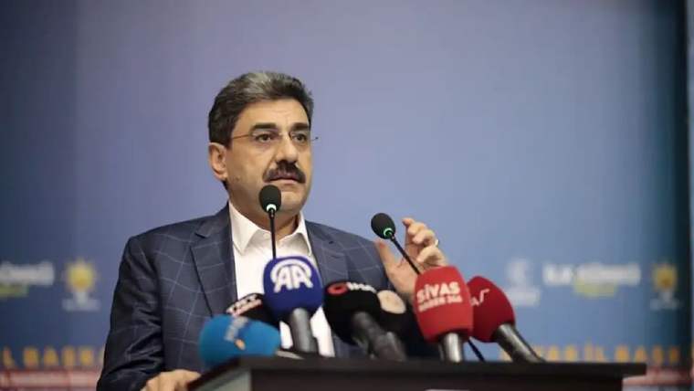 Kürsüde konuşan bıyıklı bir kişi, önündeki çok sayıda logolu mikrofon arasında elini kaldırıyor.