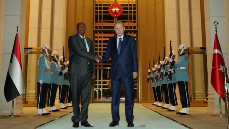 Cumhurbaşkanı Erdoğan ve el-Burhan, resmi bir törenle el sıkışıyor. Arkada Türk bayrağı ve Sudan bayrağı yer alıyor.