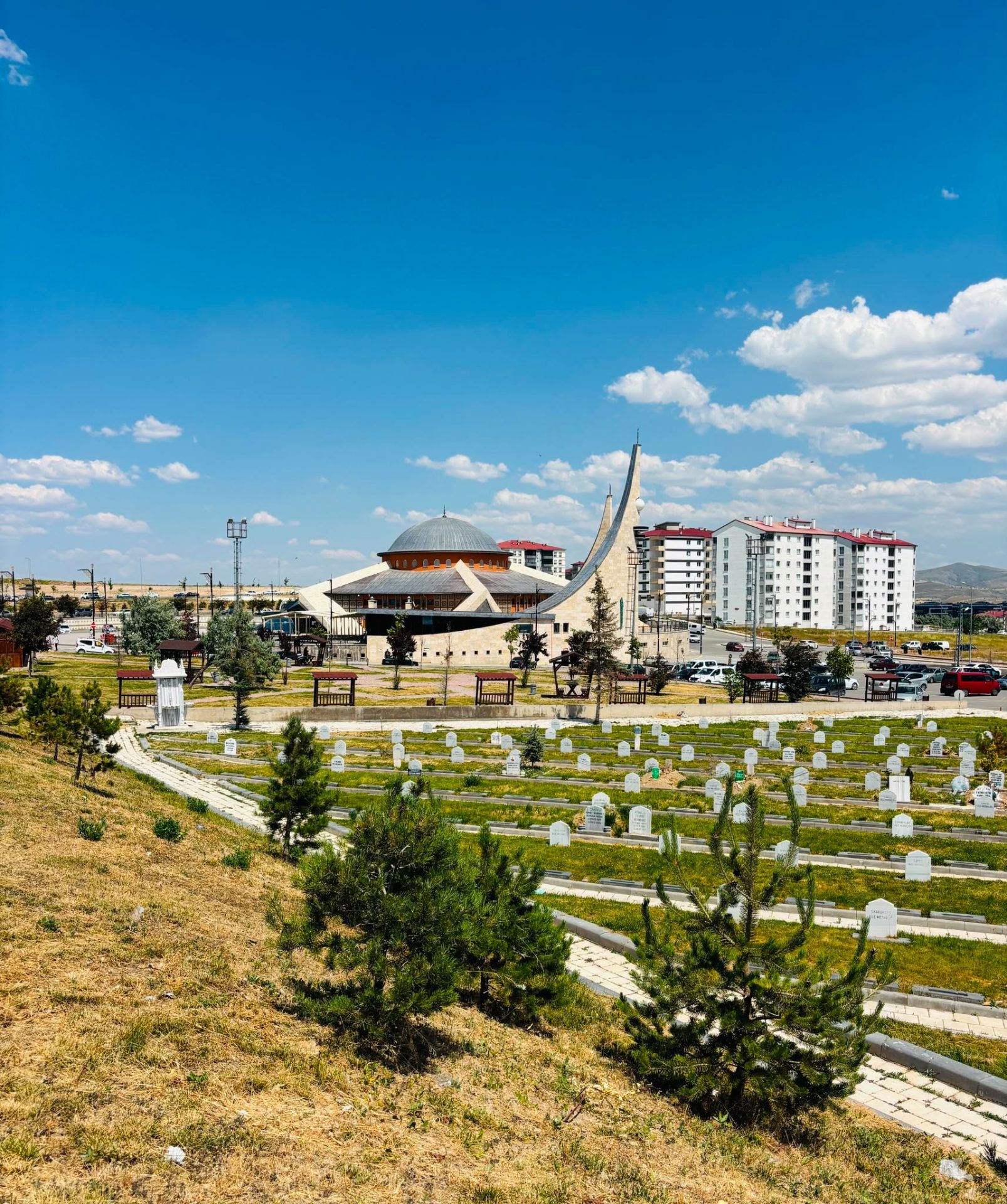 Ön planda mezar taşları bulunan mezarlık, arkasında kubbeli bir yapı ve yüksek apartmanlarla görülüyor.