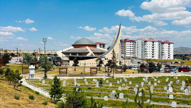 Ön planda mezar taşları bulunan mezarlık, arkasında kubbeli bir yapı ve yüksek apartmanlarla görülüyor.