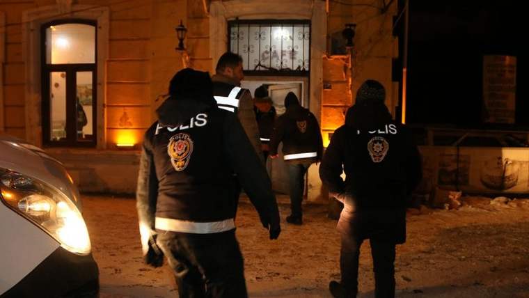 Karanlık bir sokakta, üzerinde 'POLİS' yazılı yelek giyen birkaç kişi yürüyerek bir binaya doğru ilerliyor.