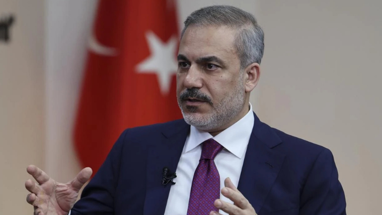 Kırmızı Türk bayrağının arkasında, konuşan bir kişi, el hareketleriyle açıklama yapıyor.