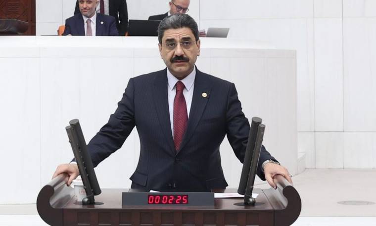 Kürsüde konuşan bıyıklı bir kişi, iki mikrofon arasında takım elbiseyle duruyor; arka planda görevliler var.