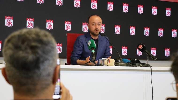 Basın masasında konuşan bir kişi, önündeki mikrofonlar ve arka plandaki kırmızı-beyaz amblemlerle görülüyor.
