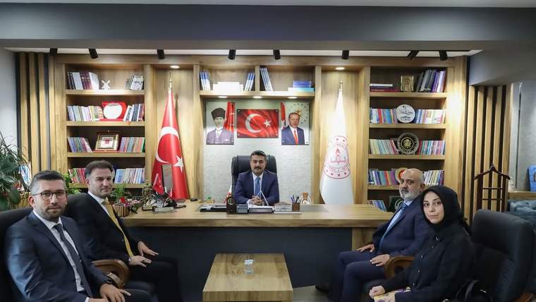 Kitaplıkların önündeki makam odasında, masa çevresinde oturan beş kişi ve Türk bayrağı görülüyor.