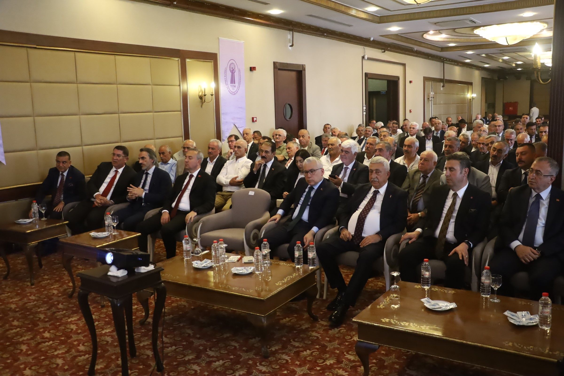 Bir toplantıda, ön sırada oturan katılımcılar dikkatle dinliyor. Arkada kalabalık bir izleyici grubu var.