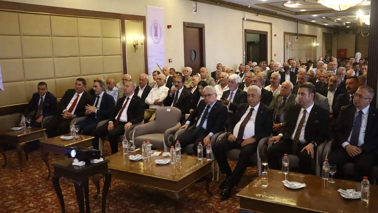 Bir toplantıda, ön sırada oturan katılımcılar dikkatle dinliyor. Arkada kalabalık bir izleyici grubu var.