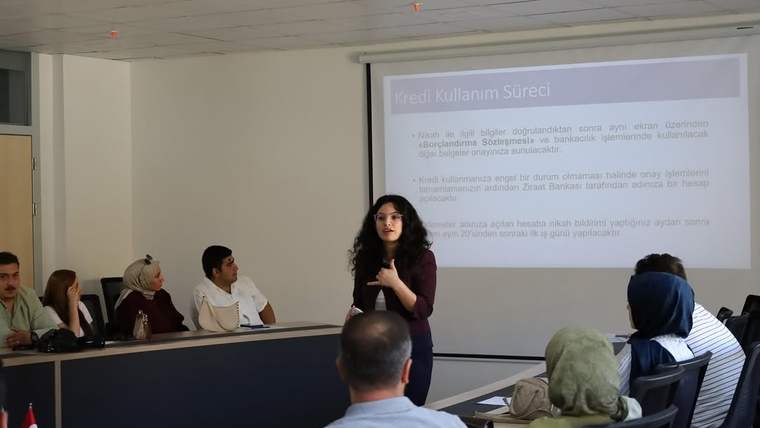 Toplantı salonunda bir kadın sunum yaparken, katılımcılar masalarda oturuyor; arkada projeksiyon perdesi bulunuyor.