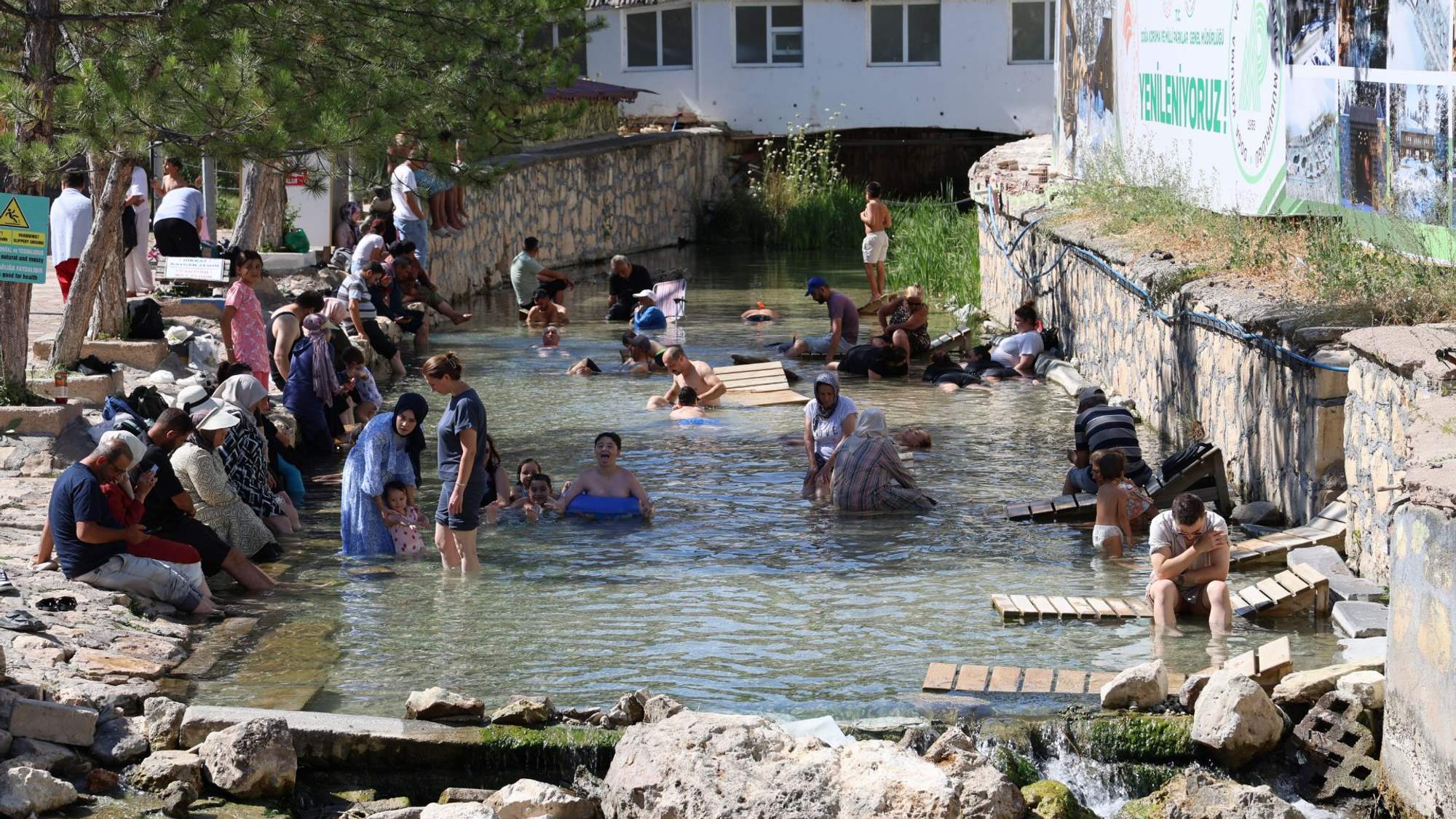 Çok sayıda kişi, taş duvarlarla çevrili sığ bir su kanalında oturuyor ve suya giriyor.